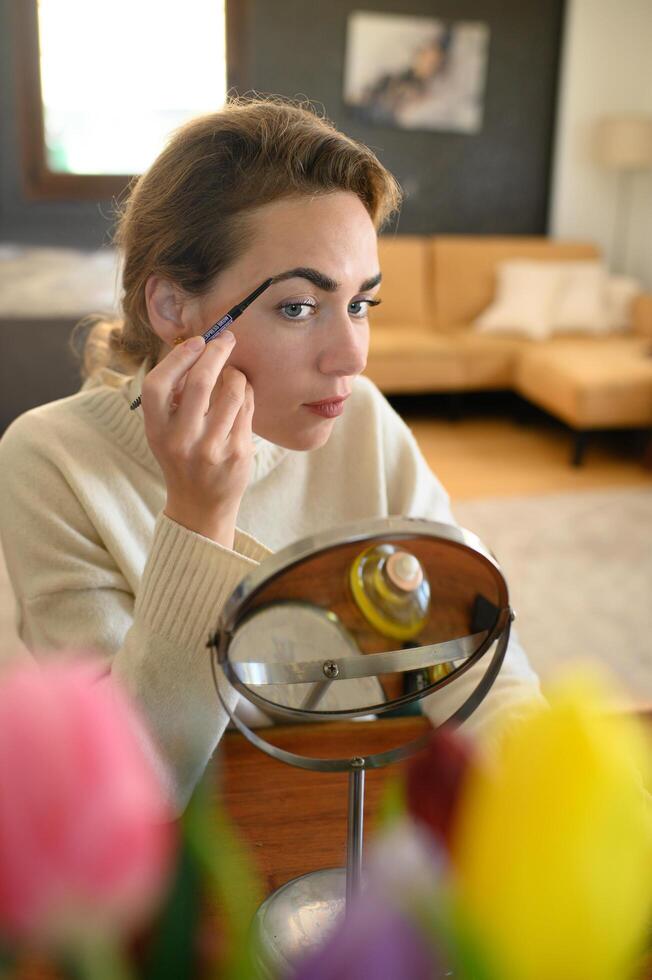 Woman shaping her brows using a brow pencil, seated at a table with makeup tools. photo