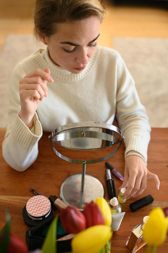 A young woman sits at a table in front of a mirror with makeup products spread out, getting ready to apply her makeup. photo