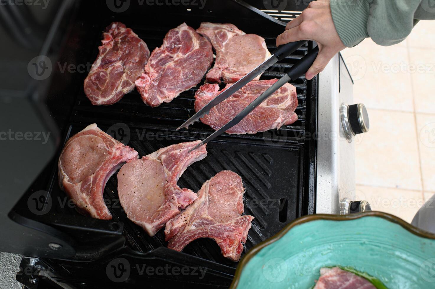Hands carefully spread fresh pork on the grill bars with tongs, marinated mushrooms visible in the background. photo