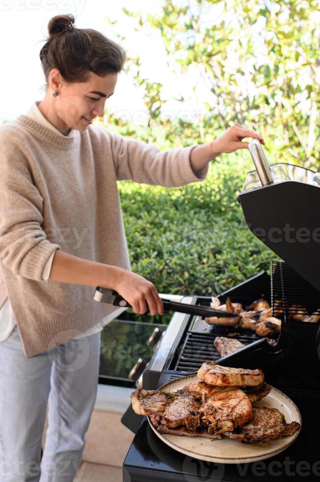 A smiling woman holds the grill steady with one hand, while using the other to place freshly grilled meat and chicken onto a plate on a beautiful terrace photo