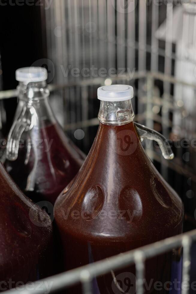 Homemade red wine in large glass demijohns with handles, sealed with plastic caps. The bottles are stored in a metal crate, showing a stage of fermentation or aging. photo