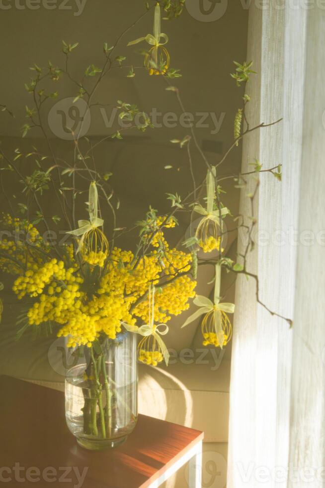 Sunny bouquet with yellow mimosa and Easter wire eggs decorations in a vase, captured in soft focus. photo