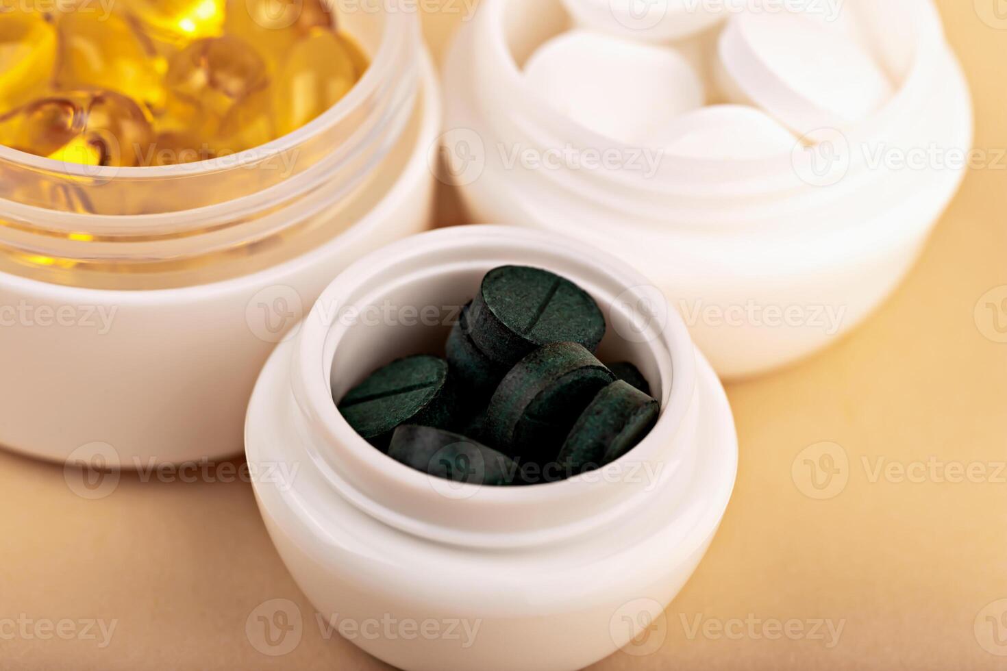 Green spirulina tablets in white cosmetic container with yellow softgel capsules and white pills blurred in background. Concept of health and dietary supplements. Closeup of spirulina tablets in jar photo