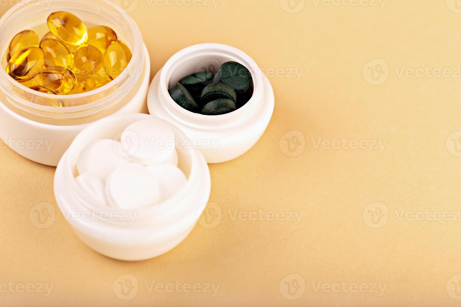 Open white jars filled with yellow capsules, green spirulina and white tablets on beige background. Concept of natural supplements and wellness routine. White containers with supplements photo