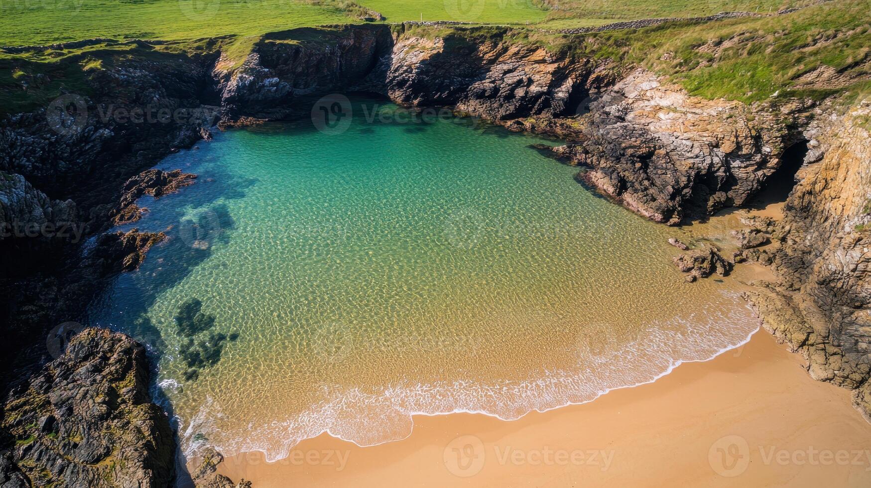 Secluded cove with clear turquoise water and sandy beach, framed by rocky cliffs and green grass. photo