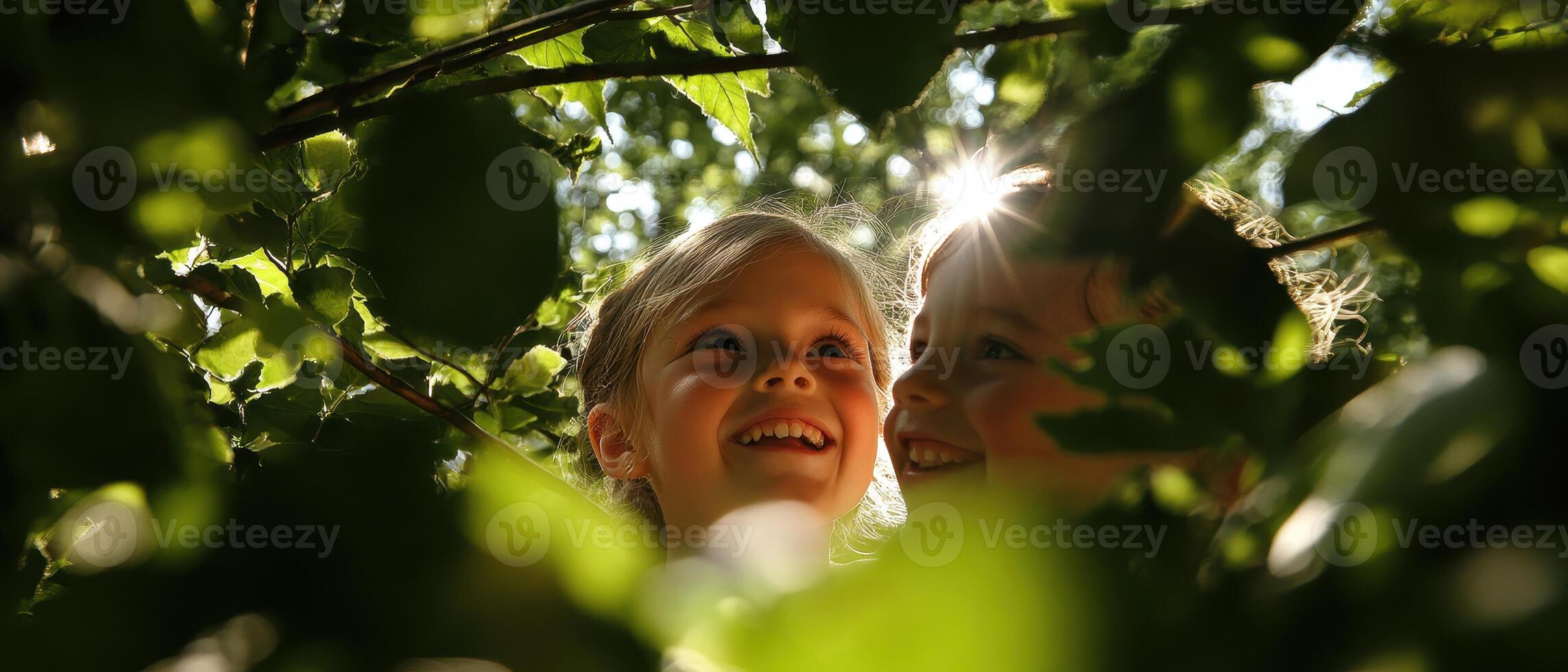 Two smiling children peek through green leaves, illuminated by sunlight, conveying joy and innocence in a natural setting. photo