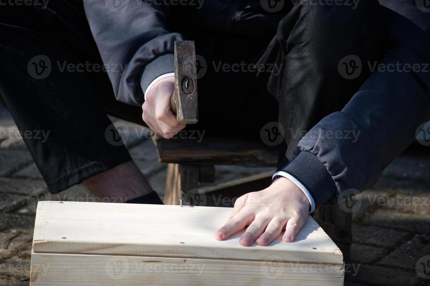 A man hammers nails into a simple wooden box he's constructing outside in the bright sunlight. photo