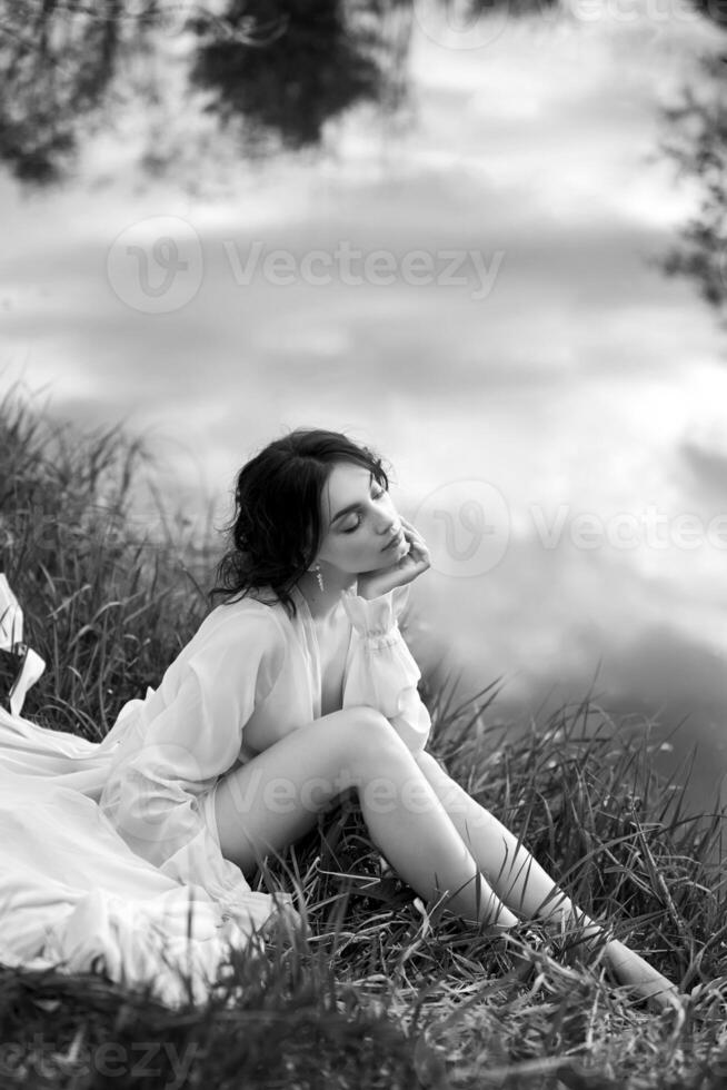 Beautiful young woman in white dress resting on shore lake pond river Professional makeup and hairstyle with curled hair photo