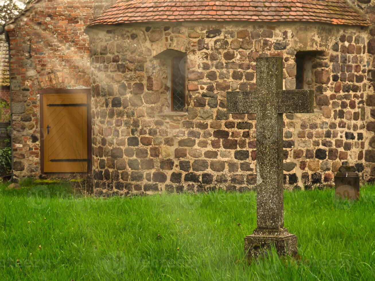 A historic stone cross in front of an old rustic church with small windows. photo