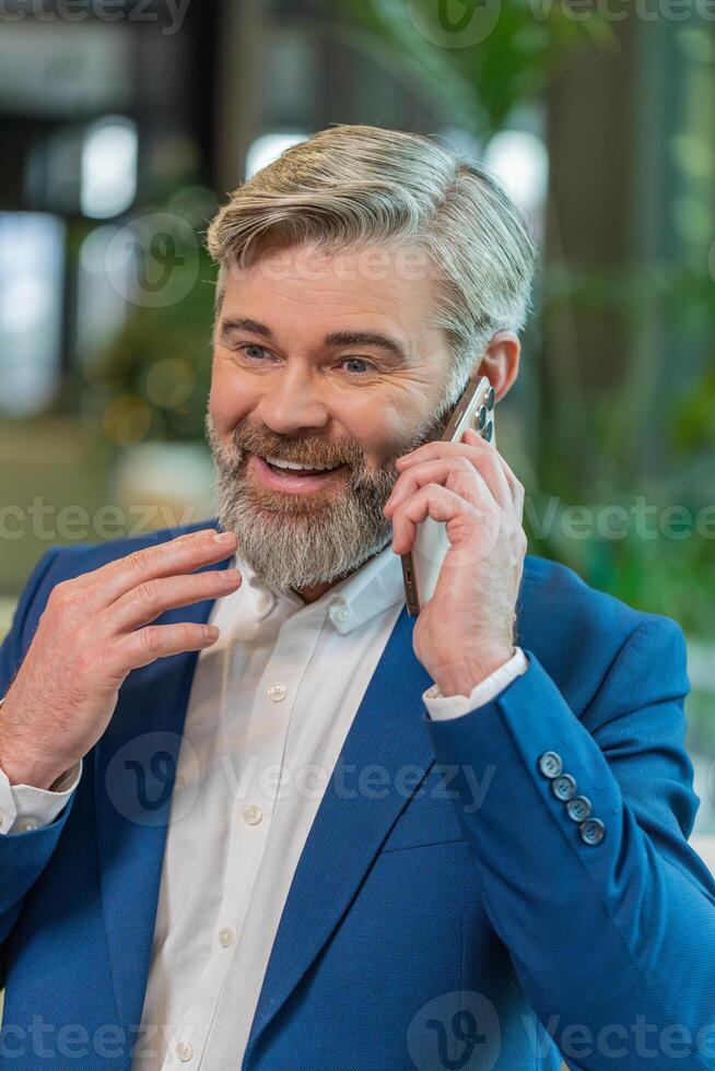 Mature businessman in formal suit answering to client on mobile phone call in modern office lobby photo