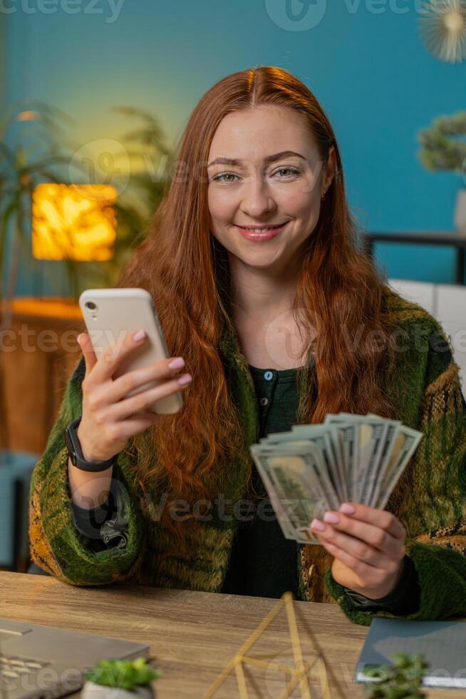 Smiling happy woman counting money dollar cash use smartphone income saves lottery win at home photo