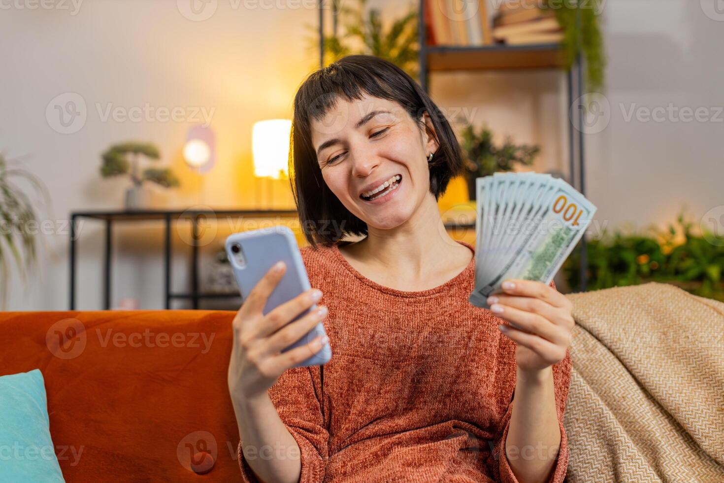 Woman counting money dollar banknotes savings earnings using smartphone calculator on sofa at home photo