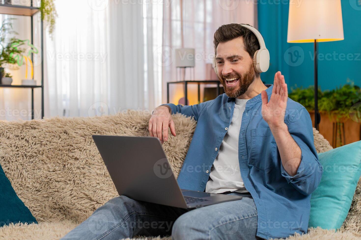 Young man in wireless headphones waving at webcam, laughing while talking on call using laptop photo
