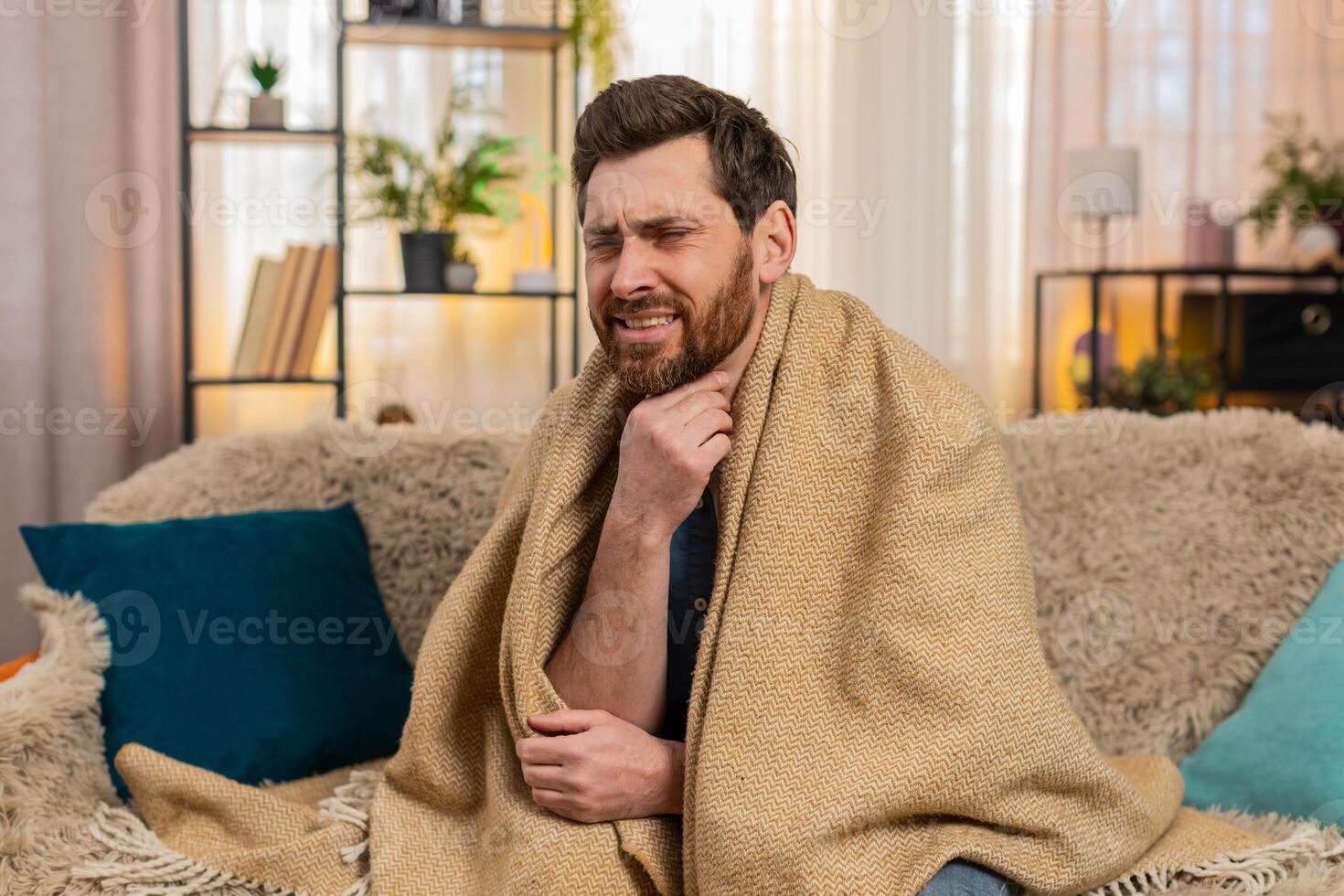 Young man sitting on the sofa at home, wrapped in a blanket, coughing, holding throat, feeling pain photo