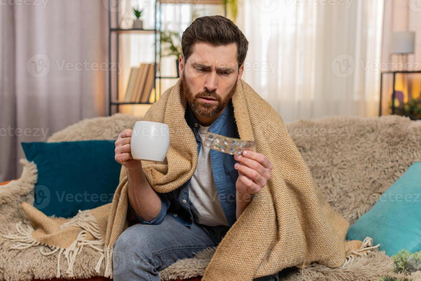Young man sitting on the sofa at home, wrapped in a blanket, holding tea, with medicine on the table photo