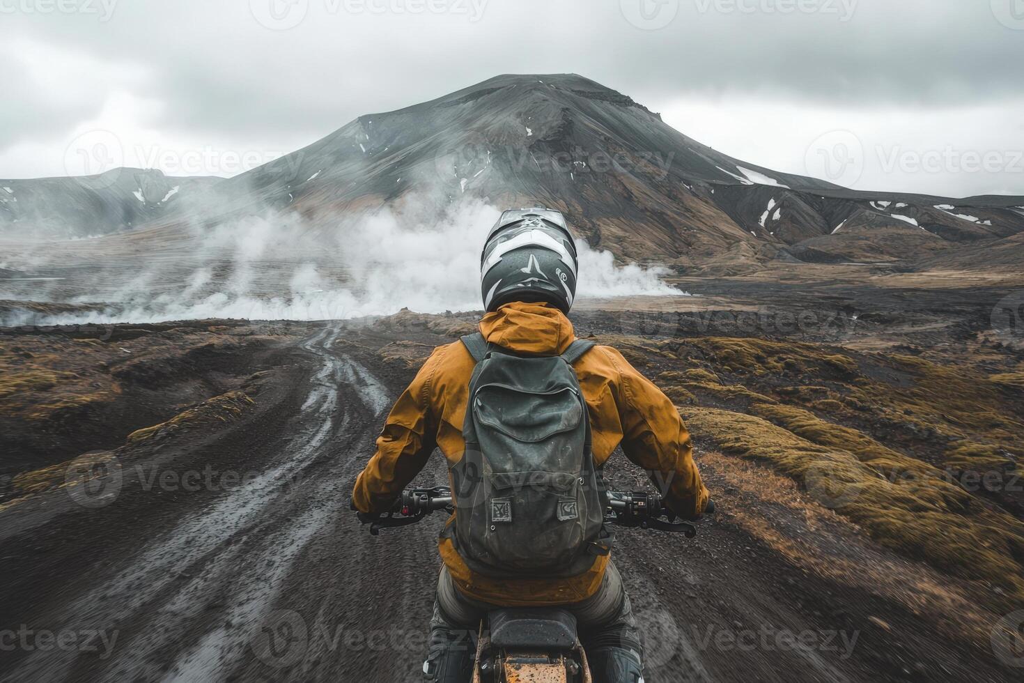 A motorcyclist wearing a helmet and a backpack navigates a muddy path across a volcanic landscape in Iceland. Steam rises from the ground, hinting at geothermal activity nearby, under an overcast sky. photo