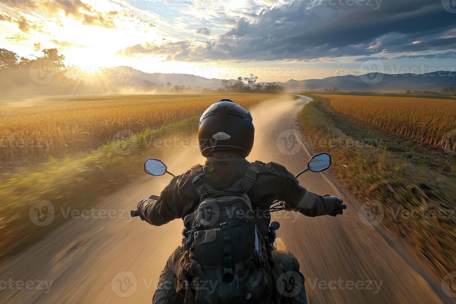 A motorcyclist navigates a winding road as the sun sets. Dust rises from the tires, creating a hazy atmosphere. Lush fields border the path, enhancing the tranquil beauty of the scene. photo