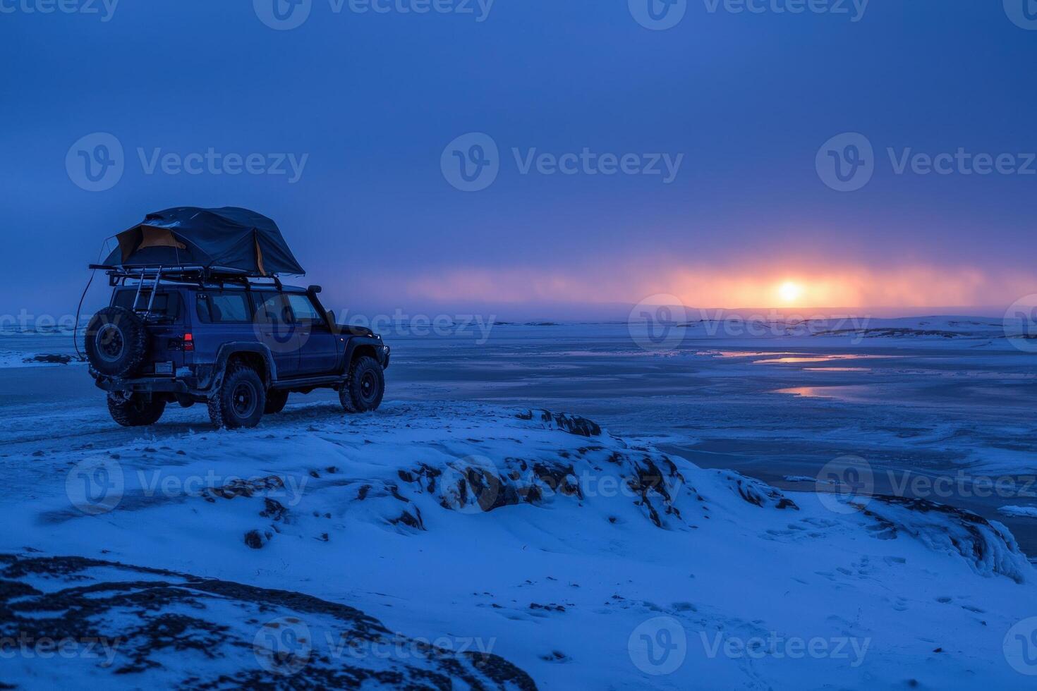 A rugged vehicle is parked on a rocky outcrop, overlooking a snowy landscape at sunset. The vibrant colors of the sky blend beautifully as night approaches in this remote winter setting. photo