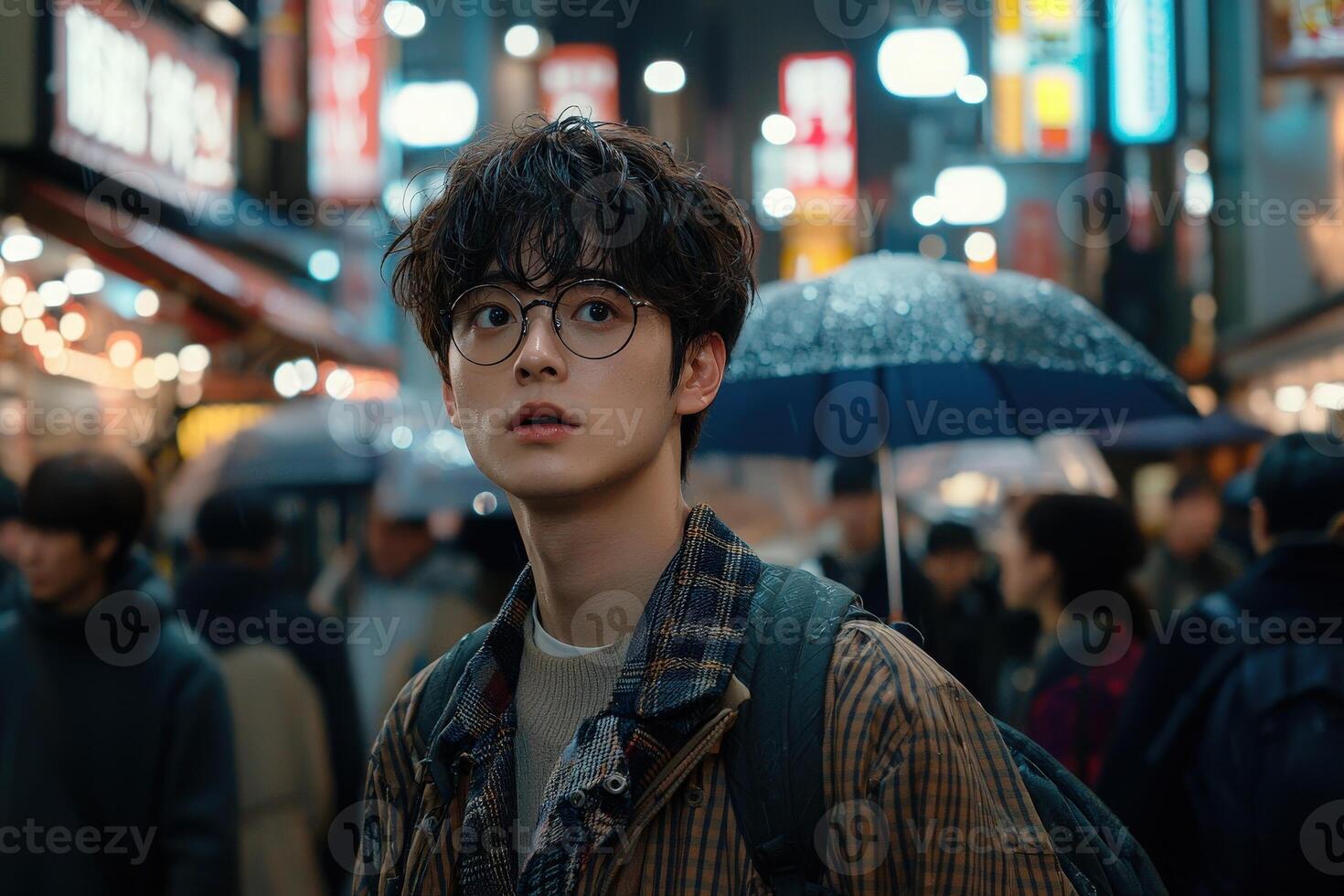 A young individual wearing glasses looks up with curiosity while navigating a bustling city street filled with bright neon signs. Rain creates a reflective surface as umbrellas pepper the crowd. photo