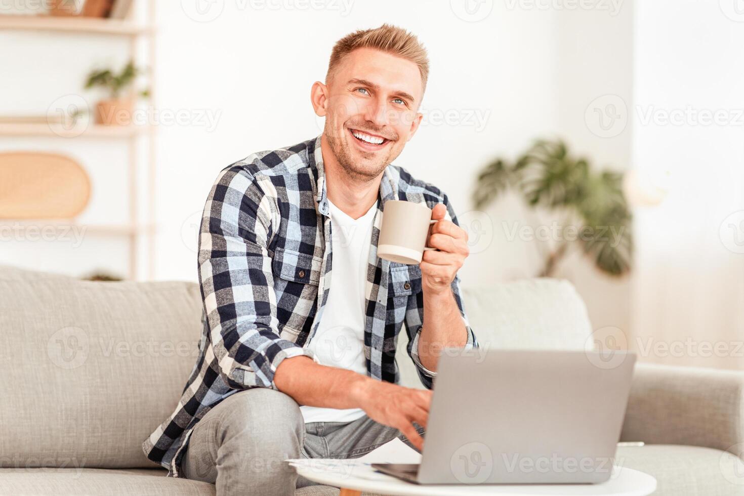 Success. Portrait of smiling confident man enjoying coffee in the morning, working on laptop in living room at home, looking up and thinking, copy space. Happy guy dreaming and holding tea cup photo
