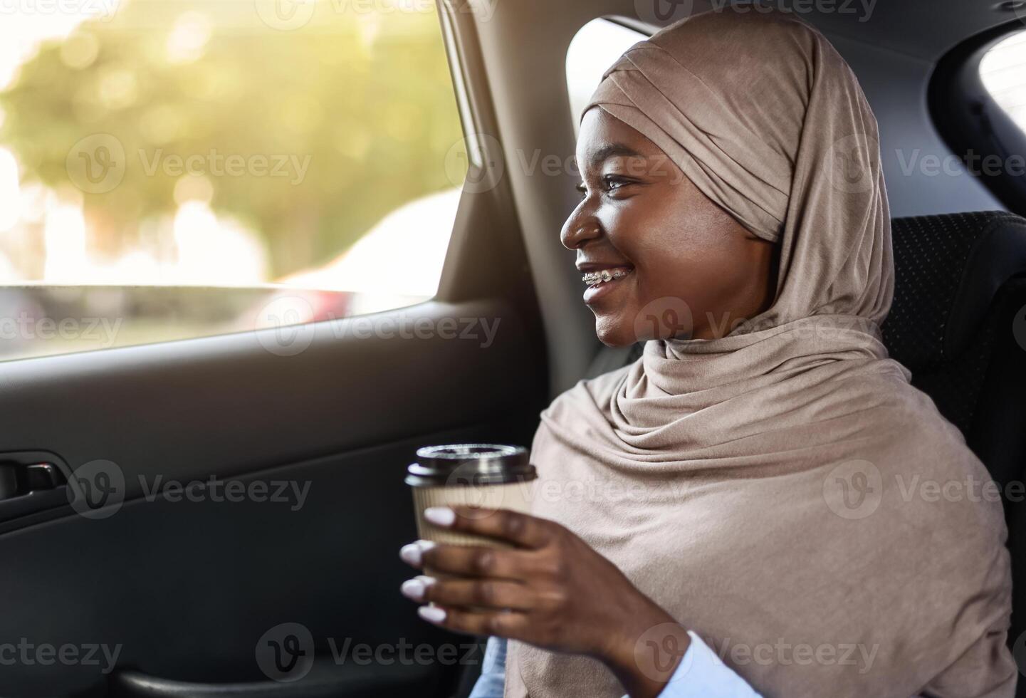 Cheerful Black Muslim Lady In Hijab Drinking Takeaway Coffee In Car On Her Way To Work, Sitting On Back Seat With Laptop And Looking From Window, Enjoying City View, Riding Taxi, Side View, Free Space photo
