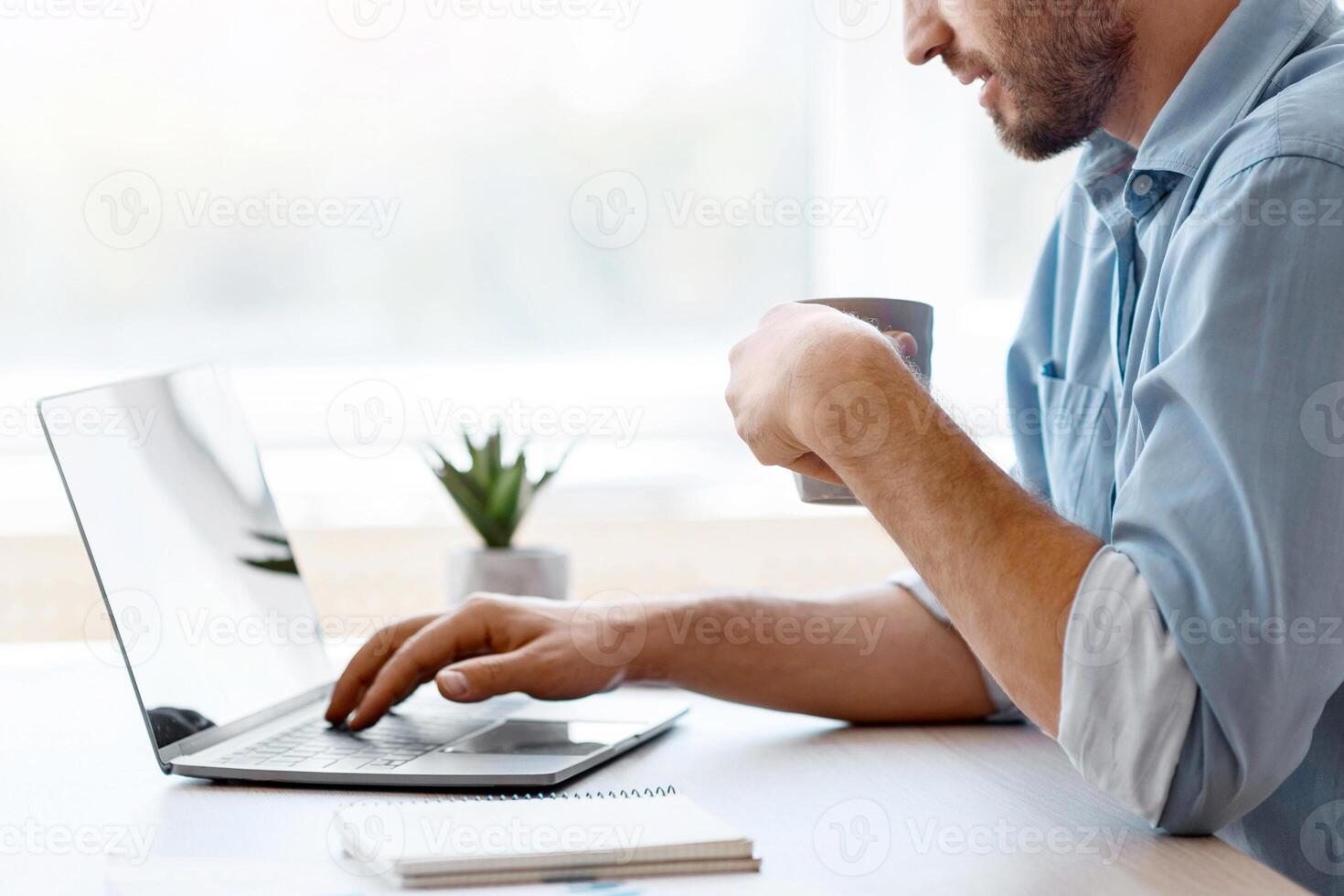 Handsome male freelancer browsing social media on laptop and drinking morning coffee, sitting at desk by the window in home office, enjoying remote work and online distance job, side view, copy space photo