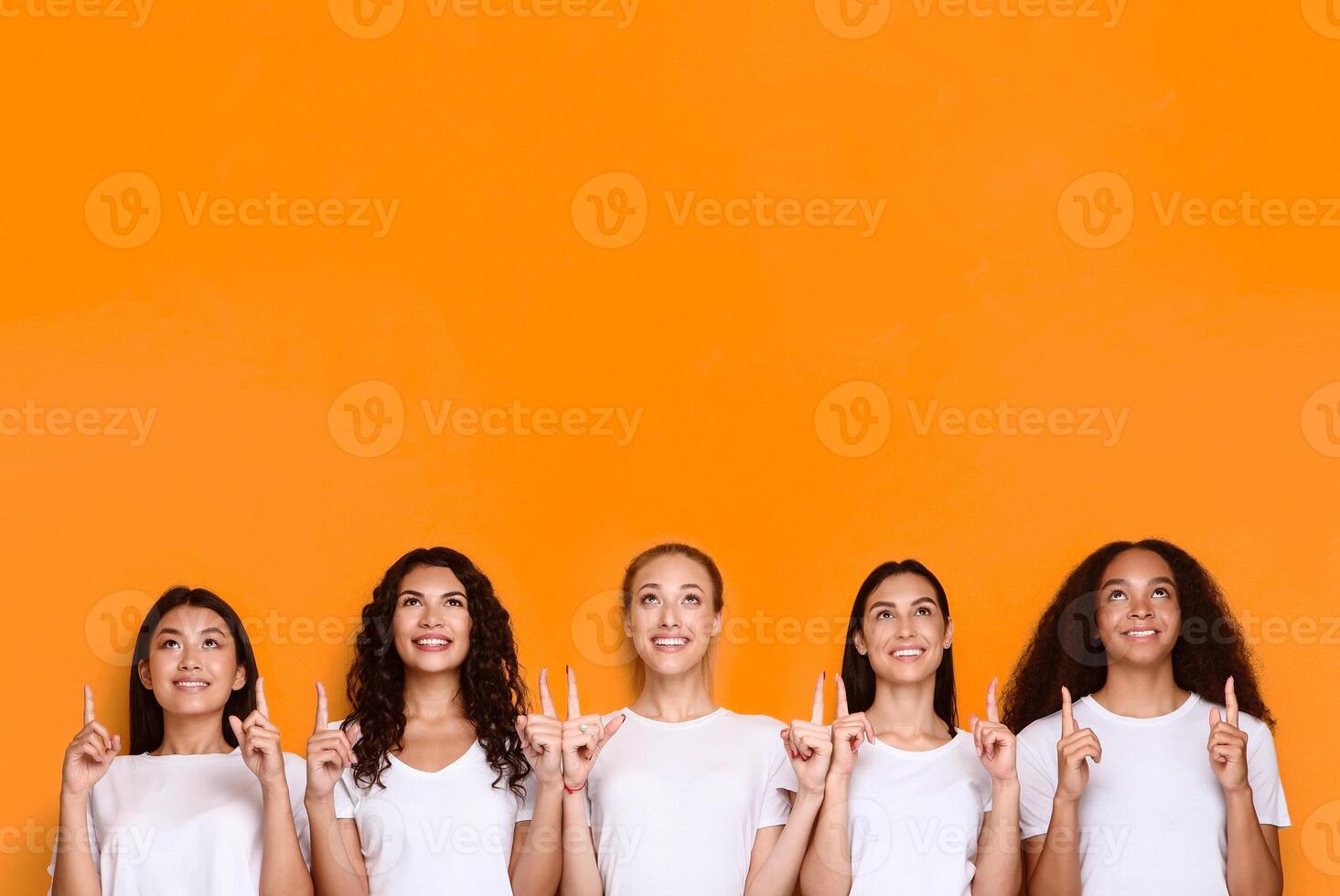 Look Upward. Five Multiethnic Women Pointing Fingers Up Posing Standing In Studio Over Yellow Background. Look There Advertisement, Females Showing Copy Space Above Head photo