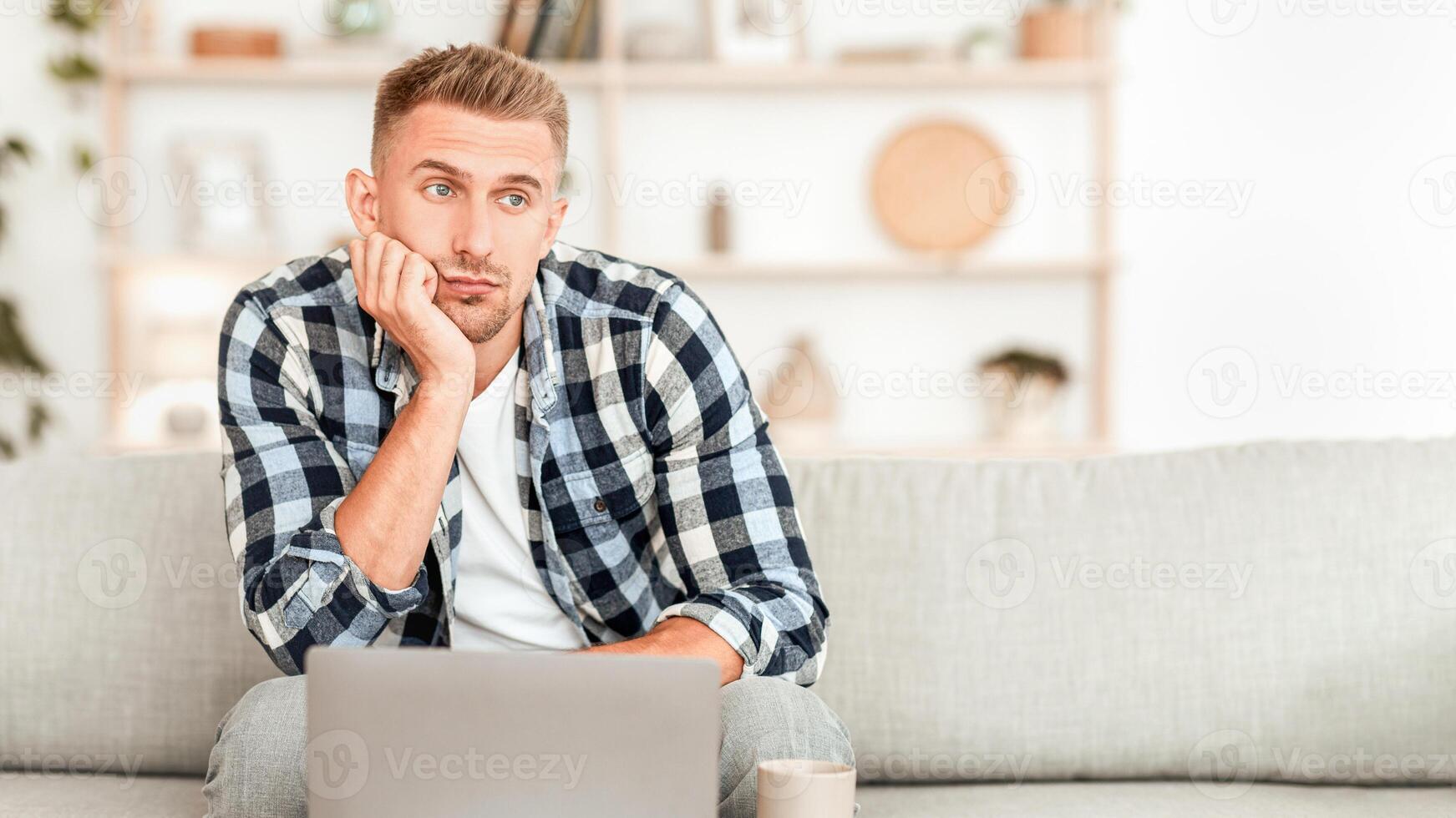 Boredom Concept. Tired man sitting on sofa with laptop, looking aside, blurred background photo