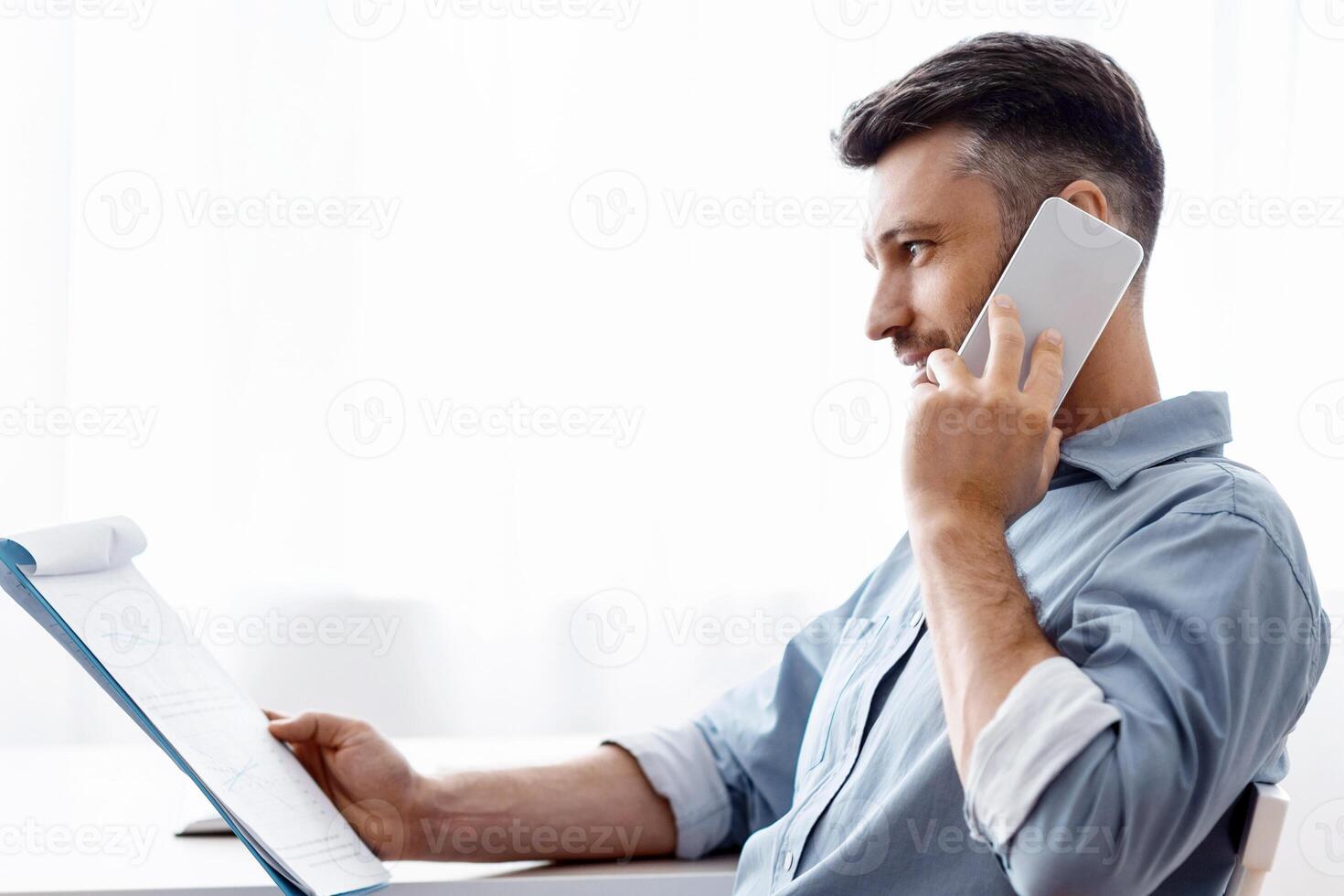 Busy Male Entrepreneur Talking On Cellphone And Checking Financial Reports At Office, Handsome Young Businessman Making Phone Call And Working With Documents, Sitting At Desk Near Window, Copy Space photo