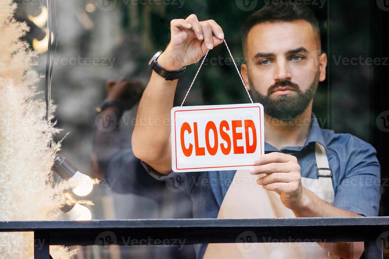 Small business, owner of flower studio closes shop at end of day and Covid-19 lockdown. Sad bearded european man seller in apron turns sign with inscription is closed in store with different plants photo