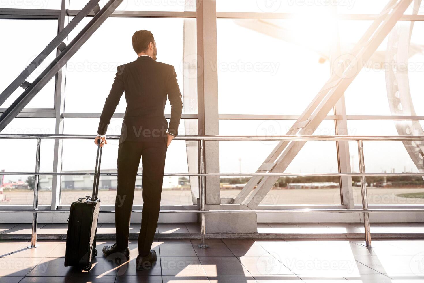 Rear view of businessman with suitcase standing next to window at airport, looking at airplane, copy space. Wealthy man in black suit waiting for departure, looking through the window, sun flare photo