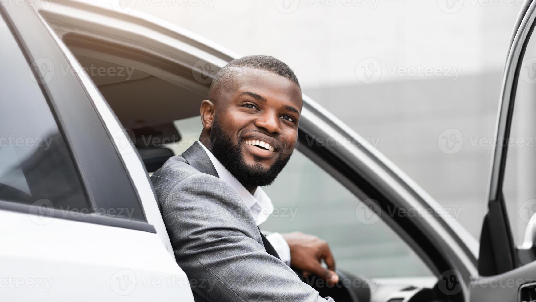 Handsome black businessman going out of the car, looking somewhere and smiling, excited about new working day. Happy african american entrepreneur arrived to office, leaving auto photo