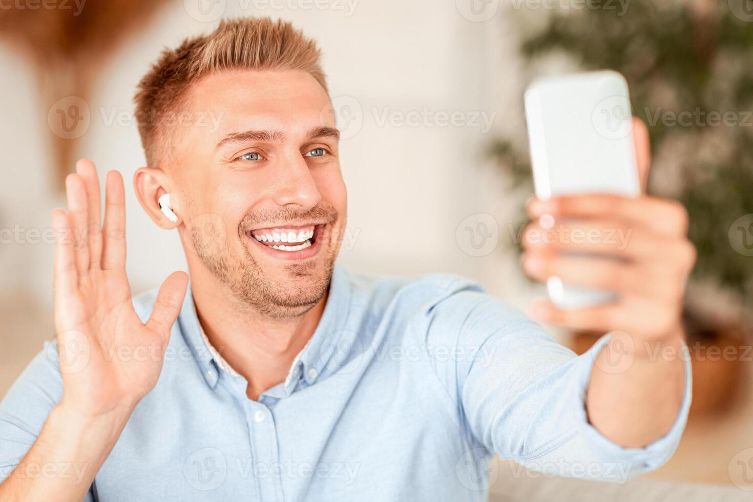 Online Call. Portrait of smiling guy making virtual conference, holding mobile phone, wearing wireless earbuds, waving with hand to webcam. Communication Concept. Man chatting using smartphone photo