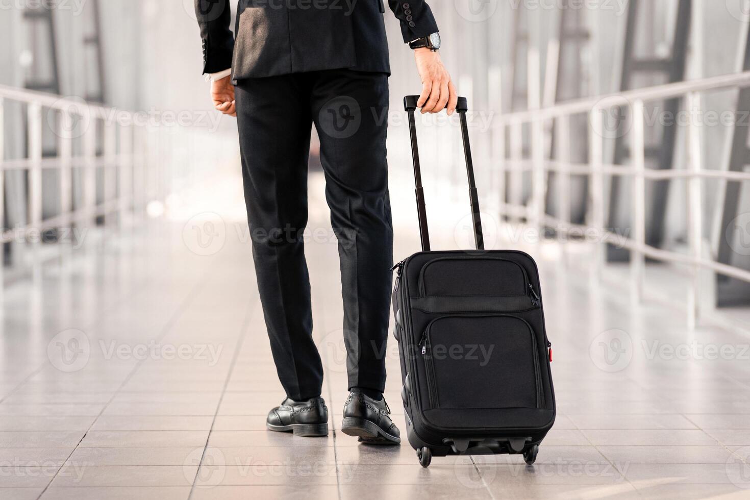 Cropped of businessman in black suit walking by airport with suitcase, back view, panorama with copy space. Unrecognizable man having business trip, going for departure or arrived for meeting photo
