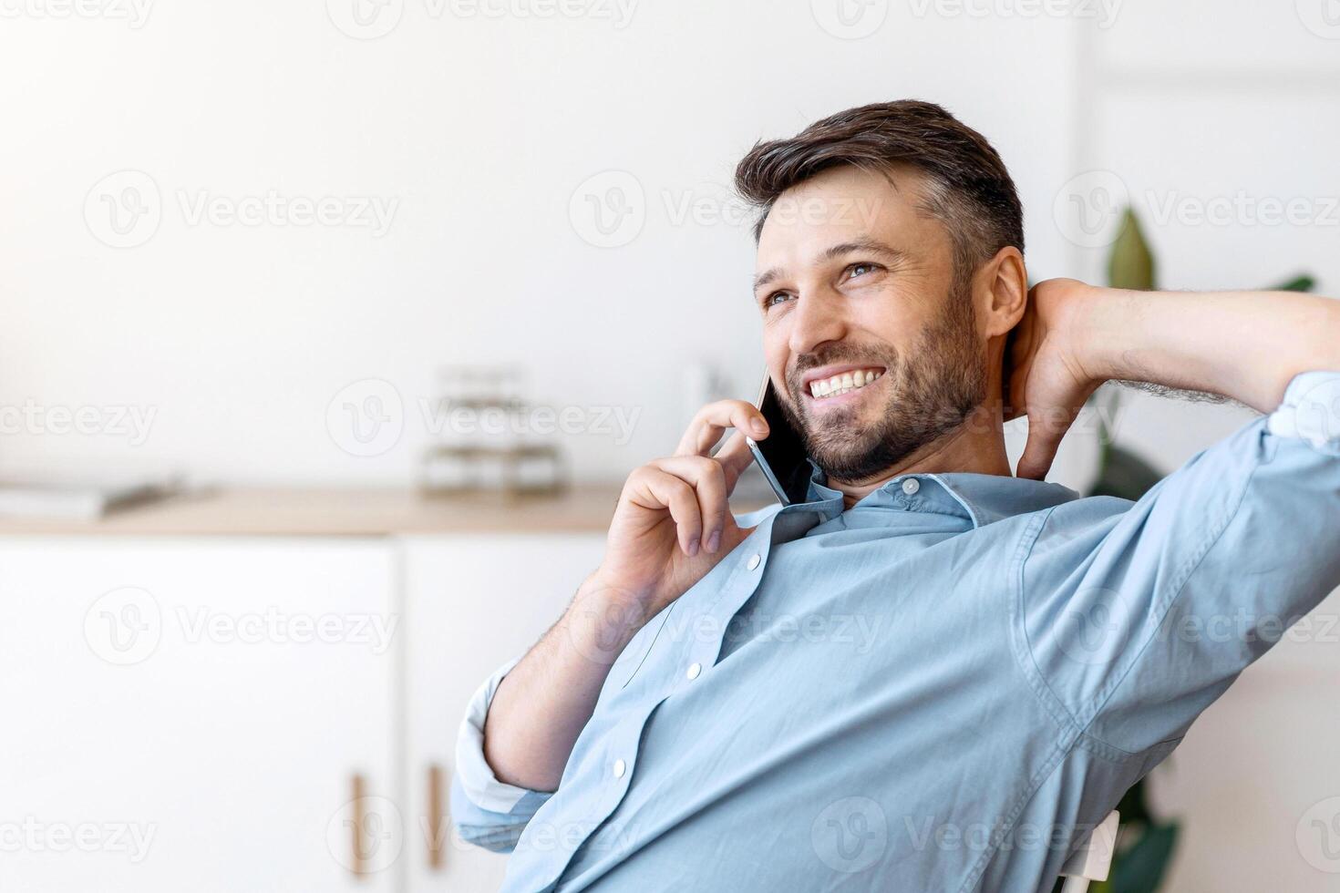 Pleasant Call. Handsome man office worker talking on cellphone and drinking coffee, relaxing at workplace, leaning back on chair, free space photo