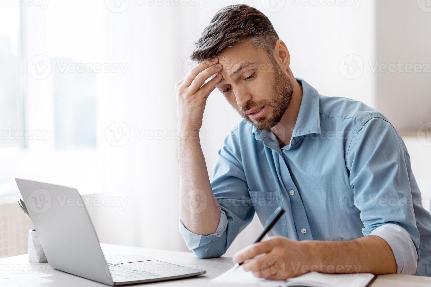 Busy Businessman Taking Notes To Notepad While Working With Laptop At Office, Planning Business Schedule, Having Deadline Stress And Problems At Work, Touching Forehead With Worried Face Expression photo
