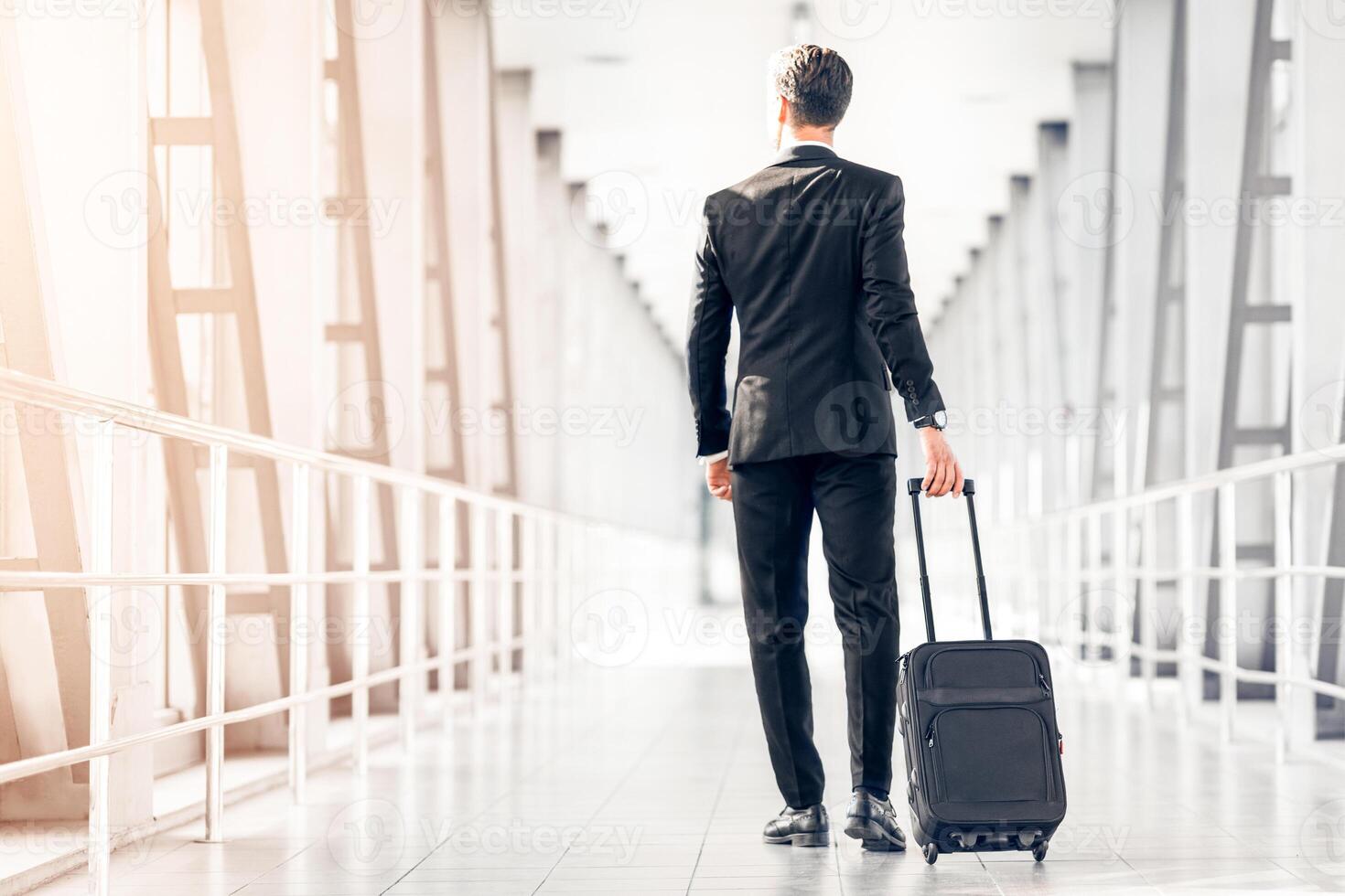 Rear view of tall businessman in stylish suit walking by airport terminal, carrying luggage, copy space. Unrecognizable entrepreneur arrived at airport, having business conference abroad photo