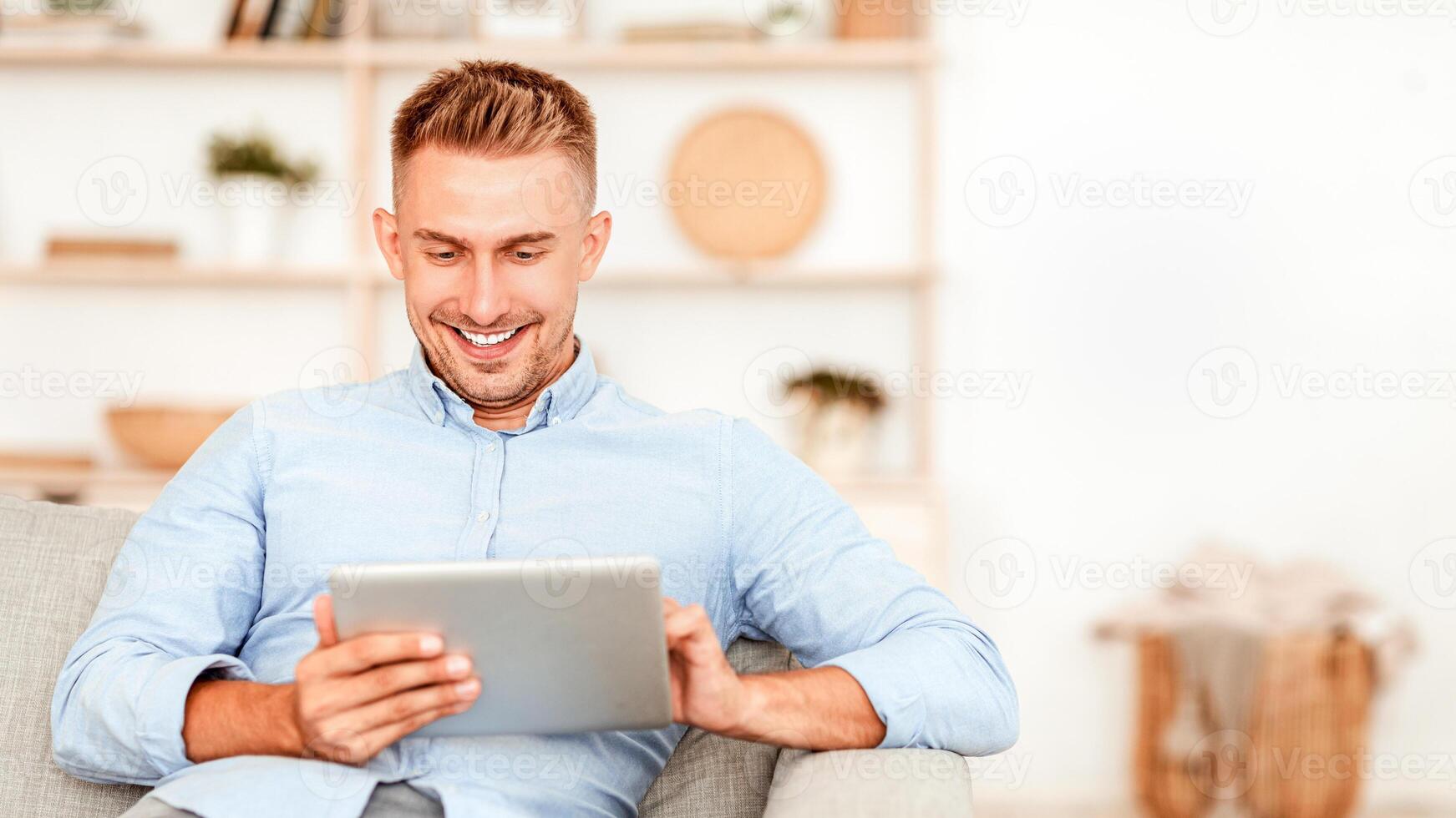 Smiling guy using digital tablet sitting on the couch at his modern apartment, copy space photo