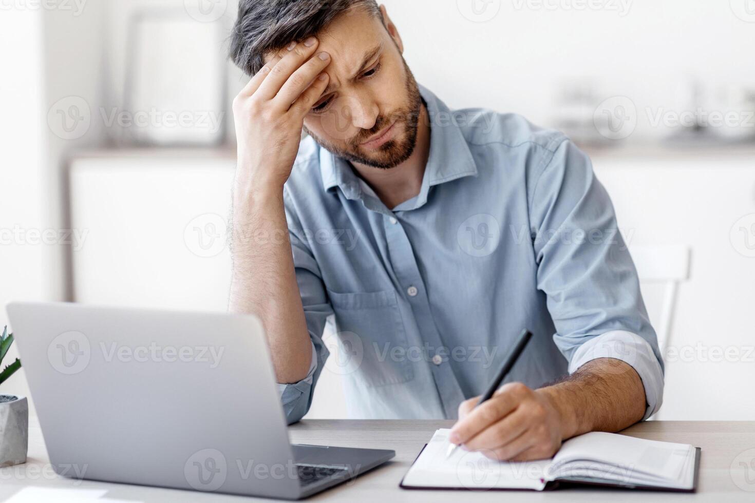 Serious Millennial Businessman Taking Notes And Working On Laptop Computer In Modern Office, Busy Entrepreneur Planning Business Schedule, Writing IN Notepad, Sitting At Desk On Workplace, Free Space photo