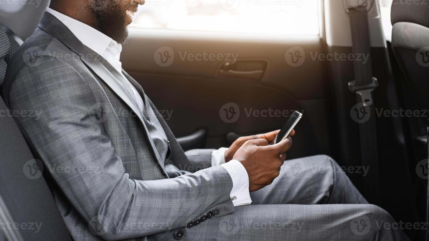 Happy african american businessman using smartphone while sitting in taxi, going to business meeting. Black guy in expensive suit chatting with business partners, going home by car, free space photo