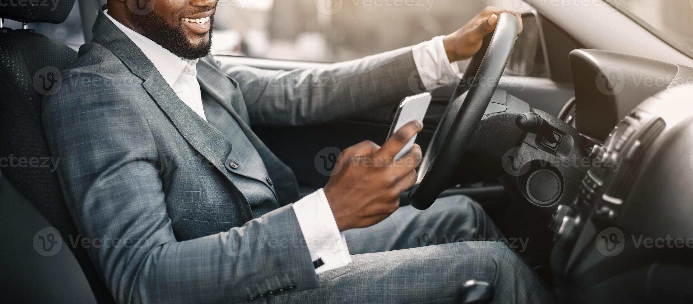Happy black guy in suit using smartphone while driving car. Positive african american businessman chatting with friends or clients on mobile phone while going to office by car, copy space photo