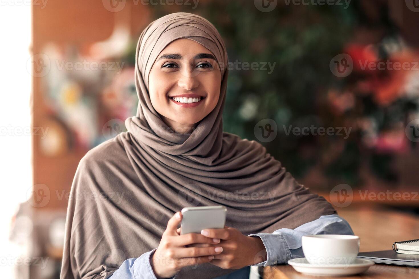 Smiling arab woman in hijab using smartphone while resting at cafe, copy space. Cheerful young arab lady drinking coffee and chatting with friends or checking social media, holding mobile phone photo