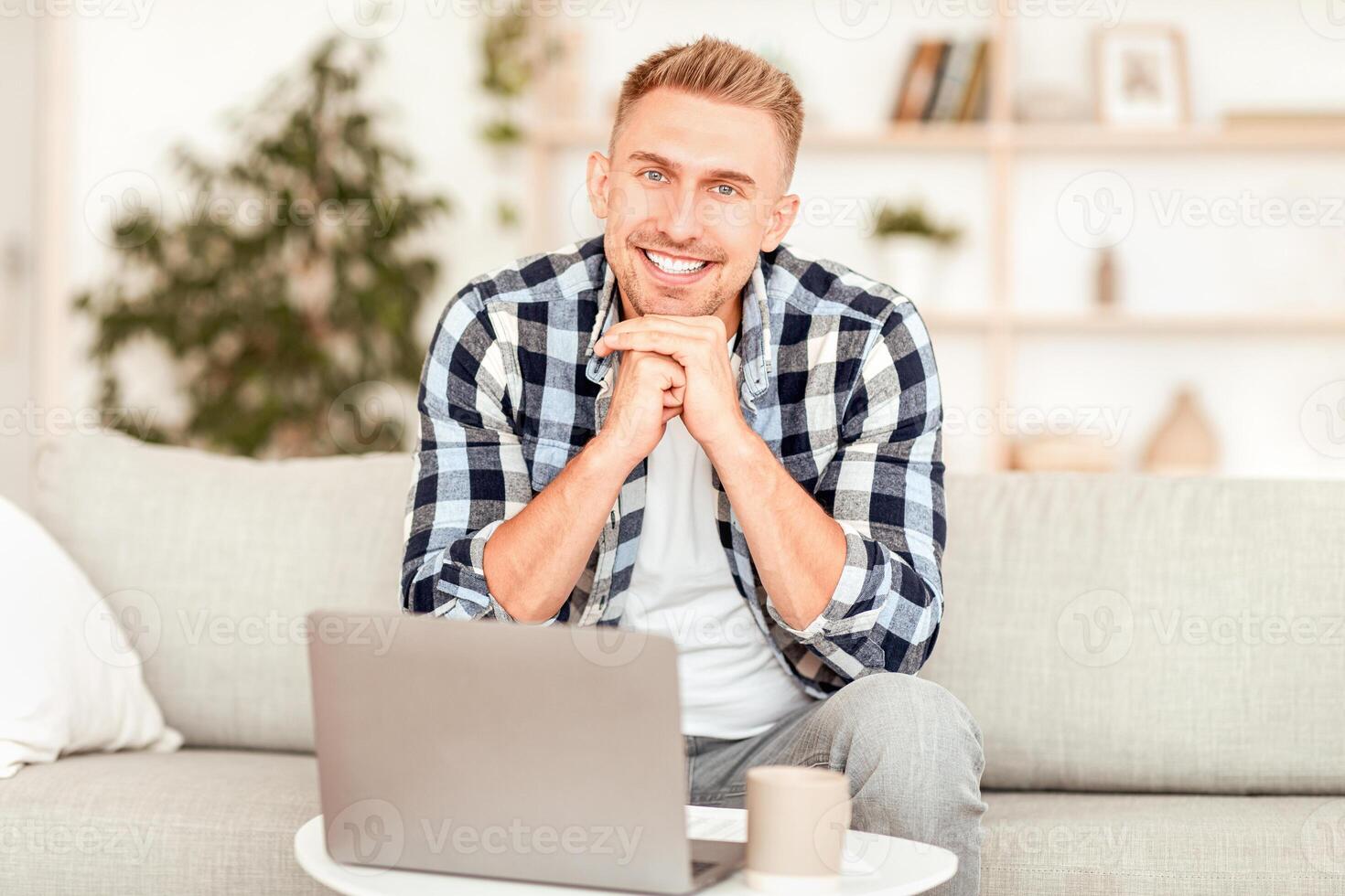Portrait Of Handsome Millennial Bearded Man In Shirt Sitting On The Couch In Living Room, Posing For Photo And Looking At Camera, Working On Laptop, Copy Space, Blurred Background, Indoors