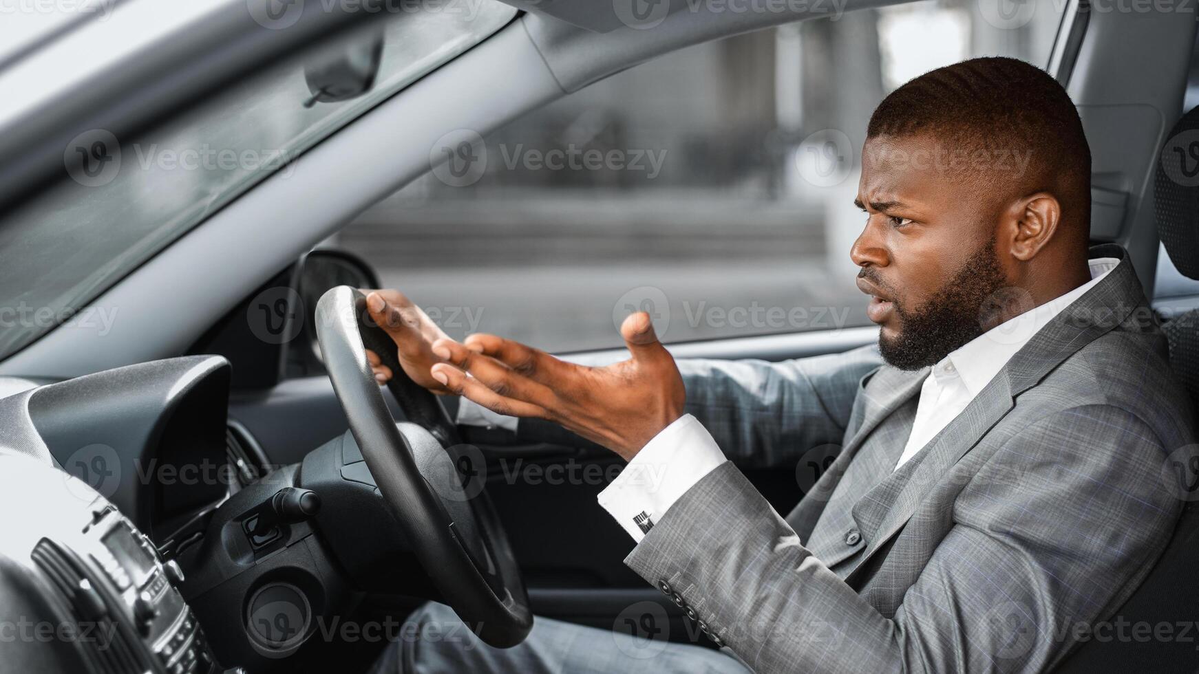 Indignant african american man driver in suit gesturing and shouting while driving car. Black businessman got stuck in traffic on the way to important business meeting, shot from inside car salon photo