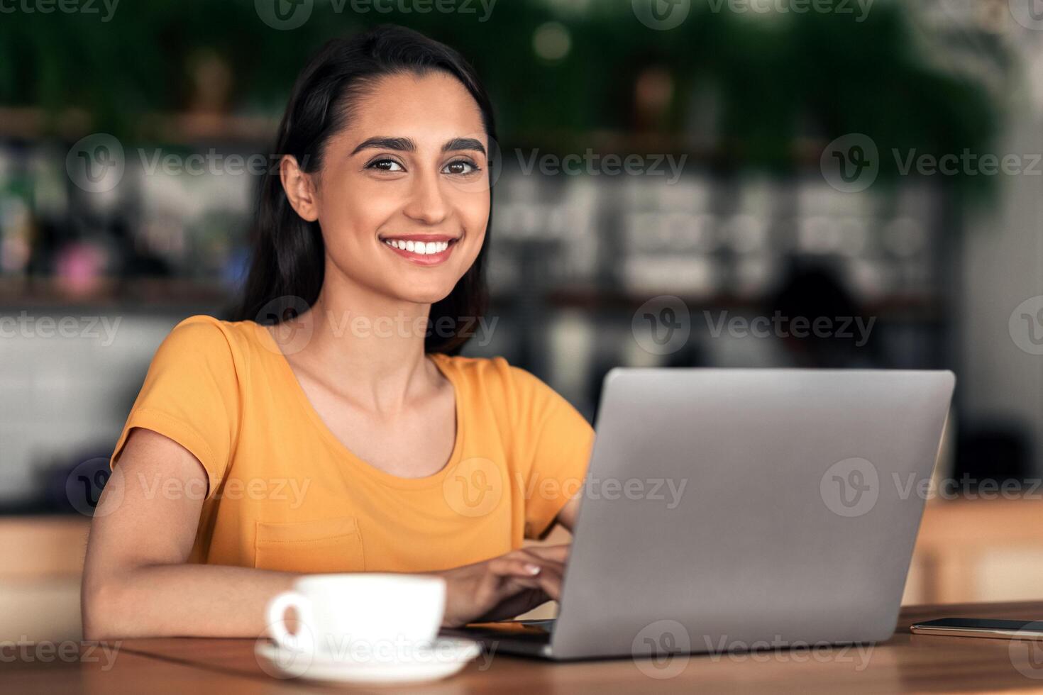 Cheerful female student using laptop at cafe, drinking coffee while studying, making report for university, copy space. Young lady sitting in front of laptop at cafeteria, smiling at camera photo