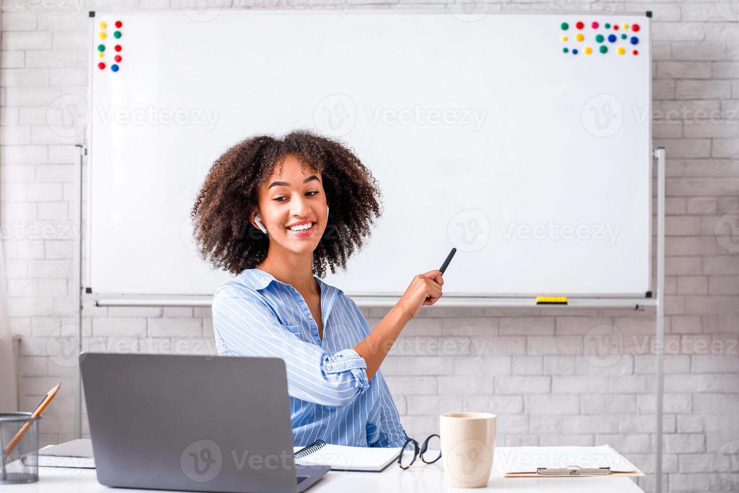Repetition of rule of English language on online lesson. Smiling african american woman points with marker at whiteboard and watch on laptop webcam sitting at table in classroom interior at home photo