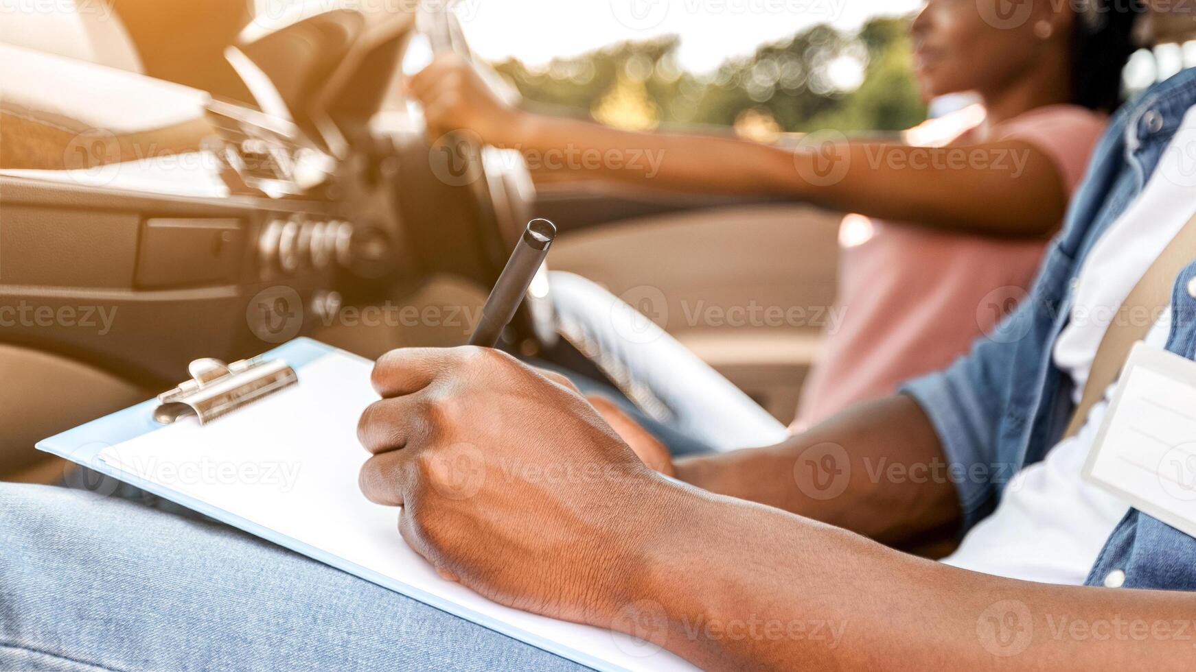 Driving school concept. Cropped of black instructor examinating young lady student, taking notes at chart while sitting by african american woman driving auto, side view, copy space photo