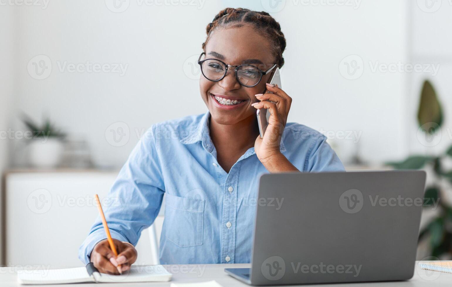 Trainee in office. Smiling black woman talking on cellphone and taking notes at workplace, young african american lady sitting at desk with laptop, having phone call and noting information, free space photo