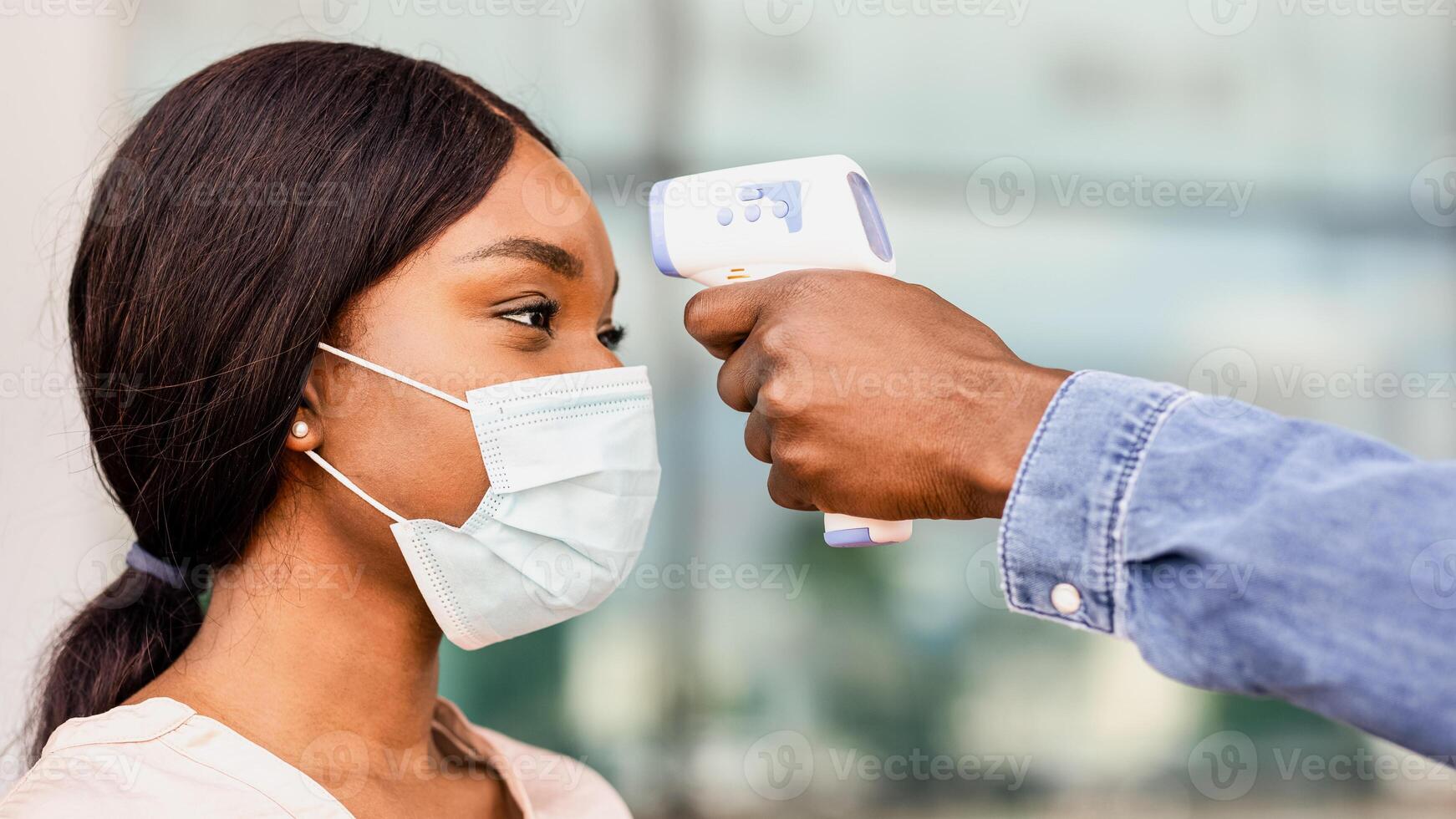 Body Temperature Control. Security guard using digital medical electronic thermometer for black woman passenger in airport due to COVID-19, closeup photo