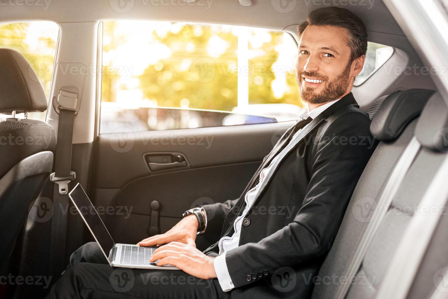 Cheerful businessman sitting in car, using laptop, working online. Handsome man in suit working on notebook while going to airport, side view. Modern technologies in business concept photo